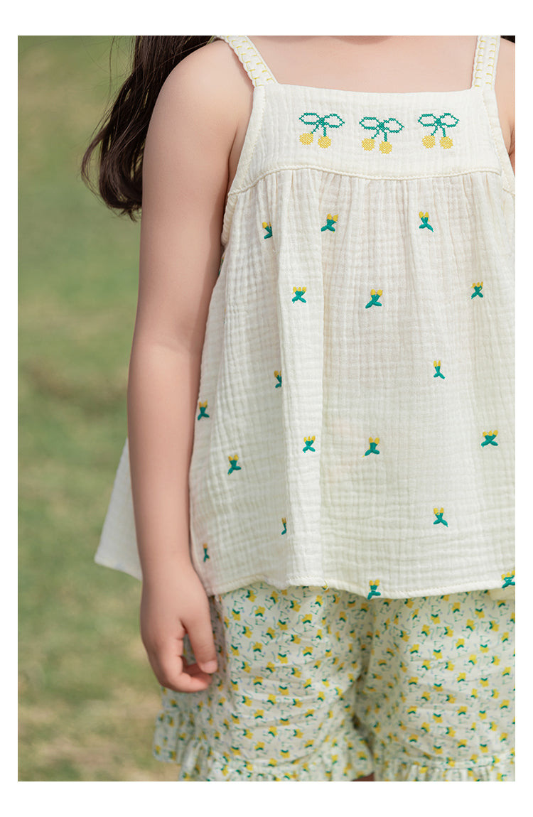 Child wearing a white sleeveless top with green and yellow cherry embroidery and floral shorts outdoors.