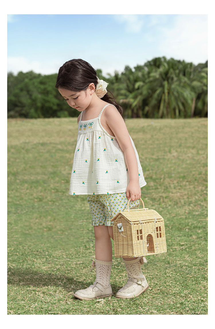Young girl in a cute sleeveless top holding a wicker basket shaped like a house in a grassy field.