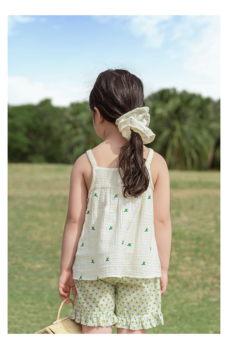 Child wearing a white cherry sleeveless top and shorts with a floral pattern, standing in a grassy field.